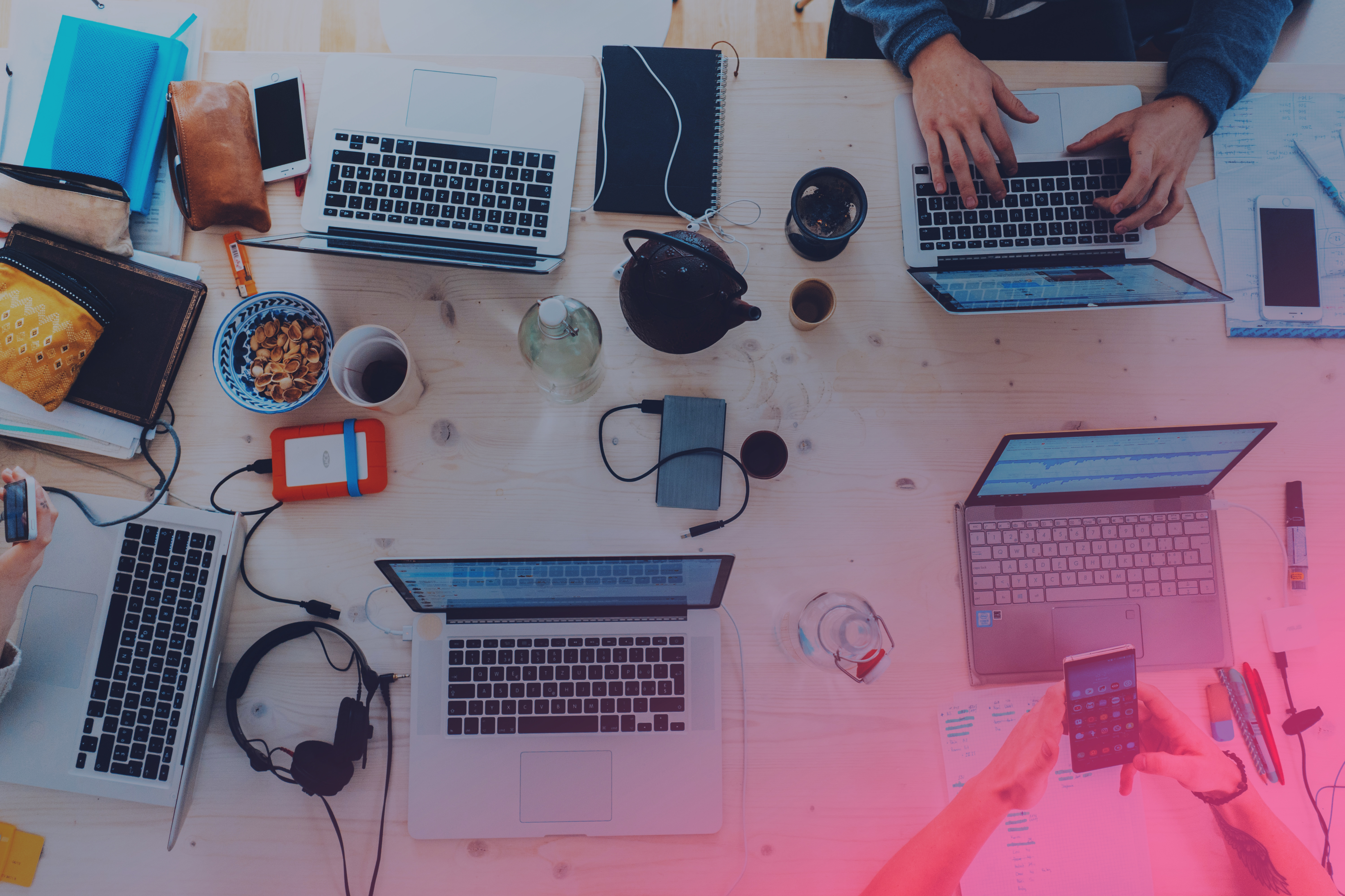 Ett skrivbord som är fyllt av datorer, sladdar, och personer som jobbar. Ger en härligt stökig känsla