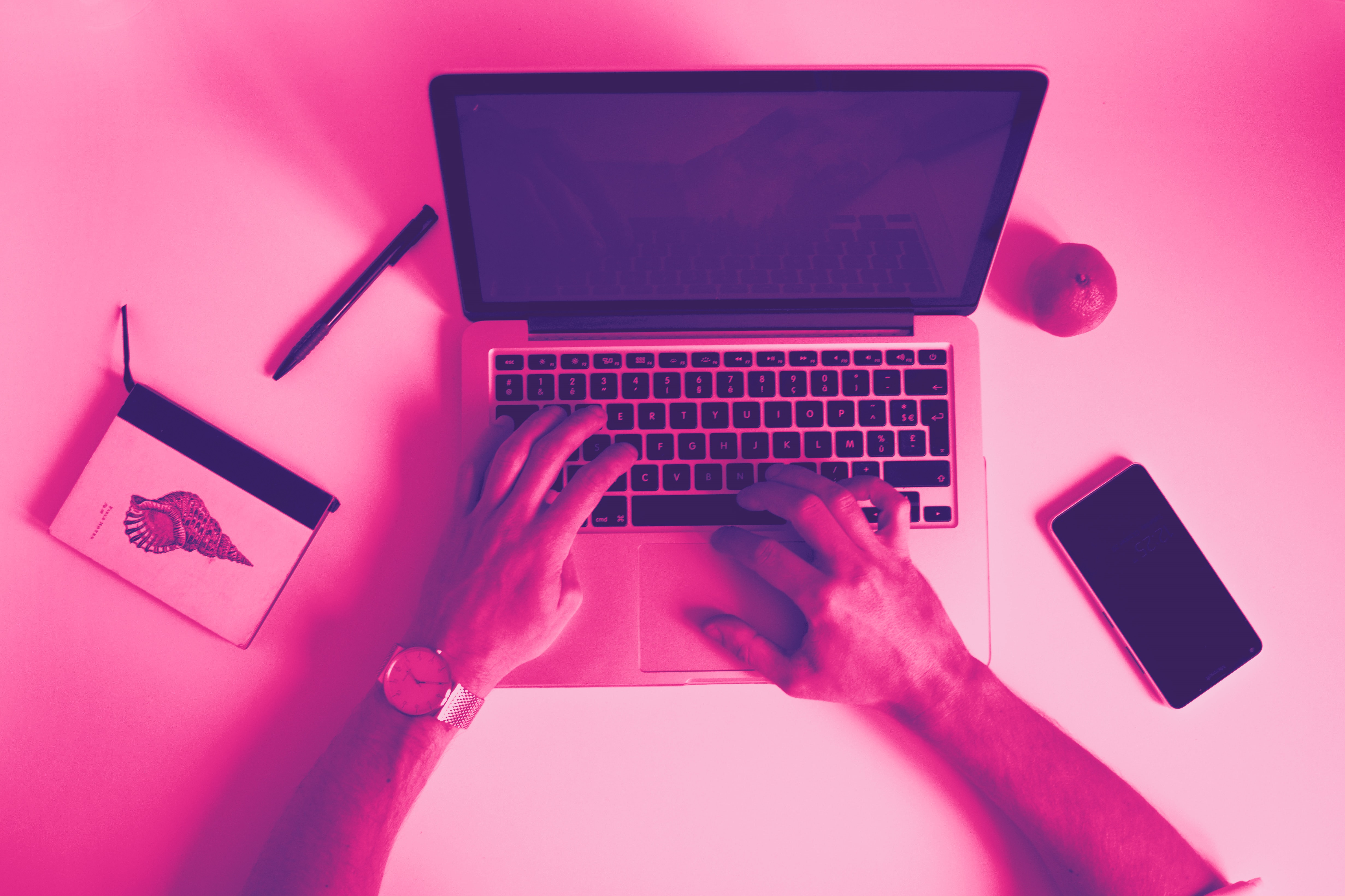 A top view of a desk. The picture shows a laptop, a notebook, mobile phone and two hands typing on the computer keyboard.