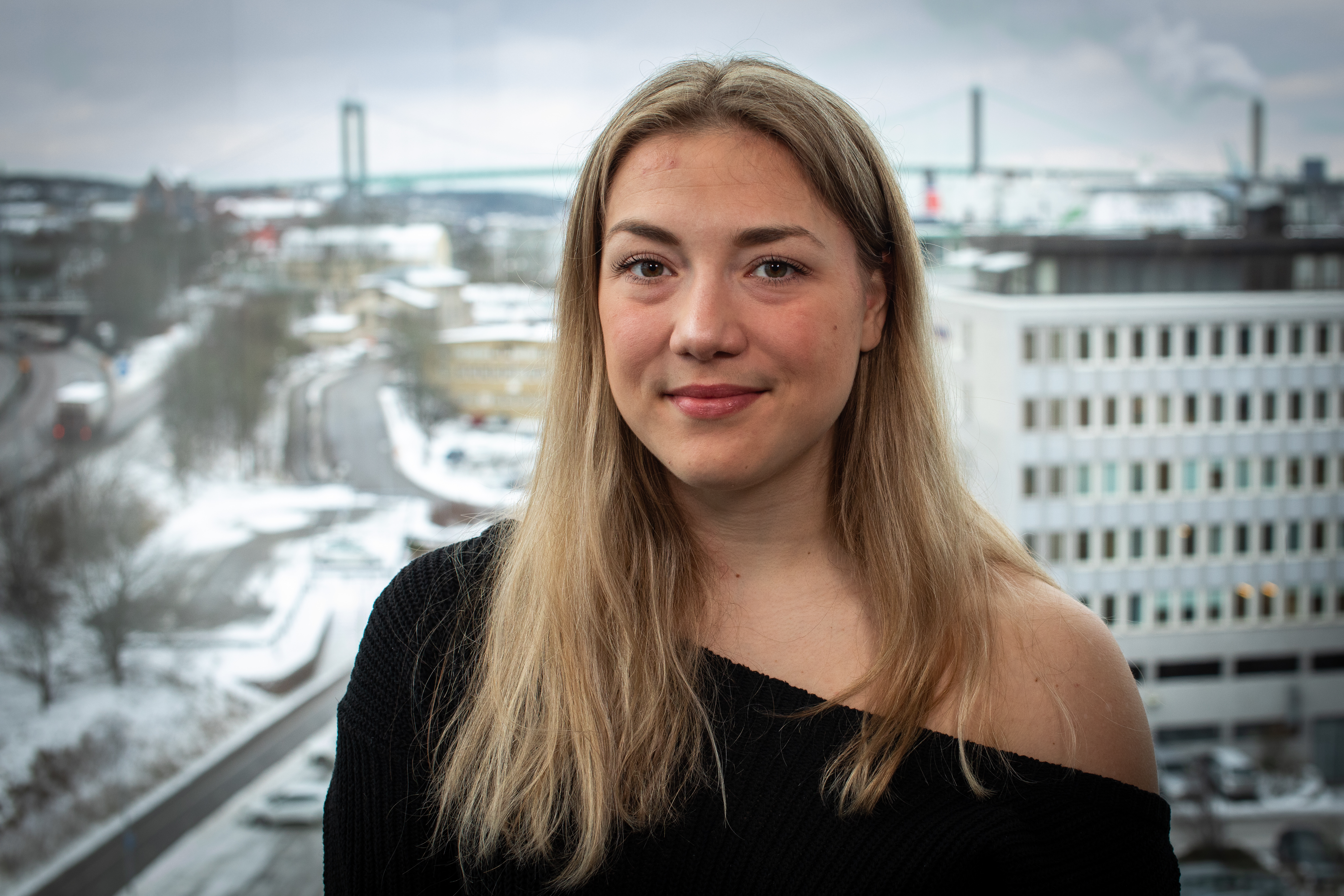 Kvinna tittar in i kameran. Hon st&aring;r framf&ouml;r ett klart f&ouml;nster och man kan sk&aring;da en storstad bakom henne.